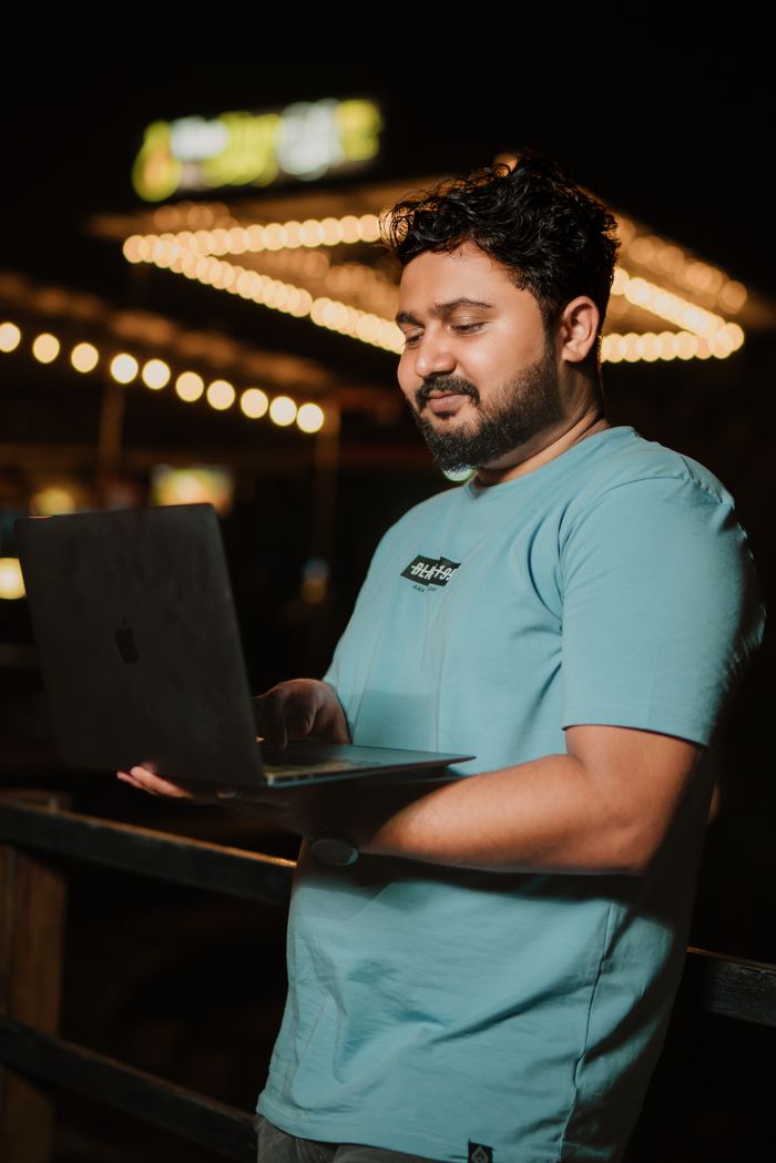 Home A man in a t-shirt using a laptop outdoors at night, with a serene ambiance and bokeh lights.