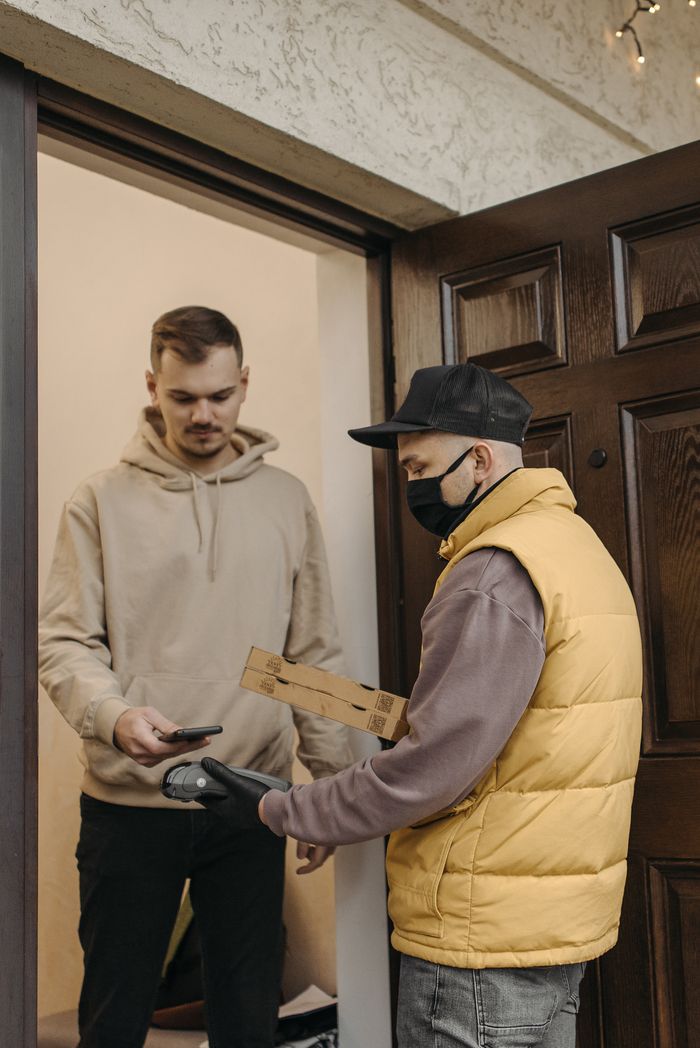 A deliveryman in a mask hands a package to a customer making a contactless payment.
